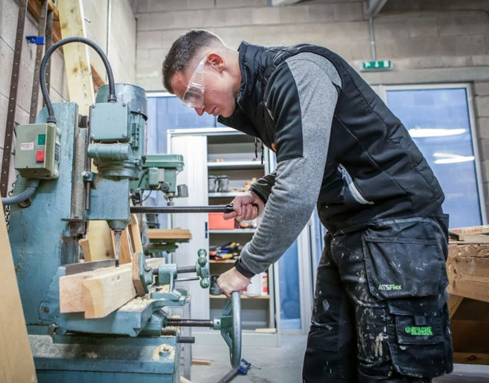 A joinery student wearing safety glasses, operating woodworking machinery to shape timber in a college workshop. A joinery student wearing safety glasses, operating woodworking machinery to shape timber in a college workshop.